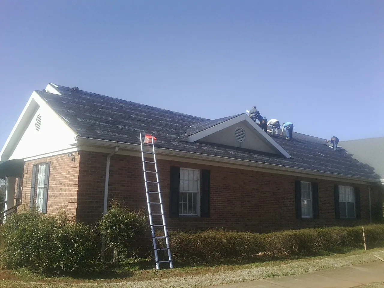Crew tearing shingles off a brick ranch roof with a ladder set — Emergency Roof Repair work by our crew in Conesus