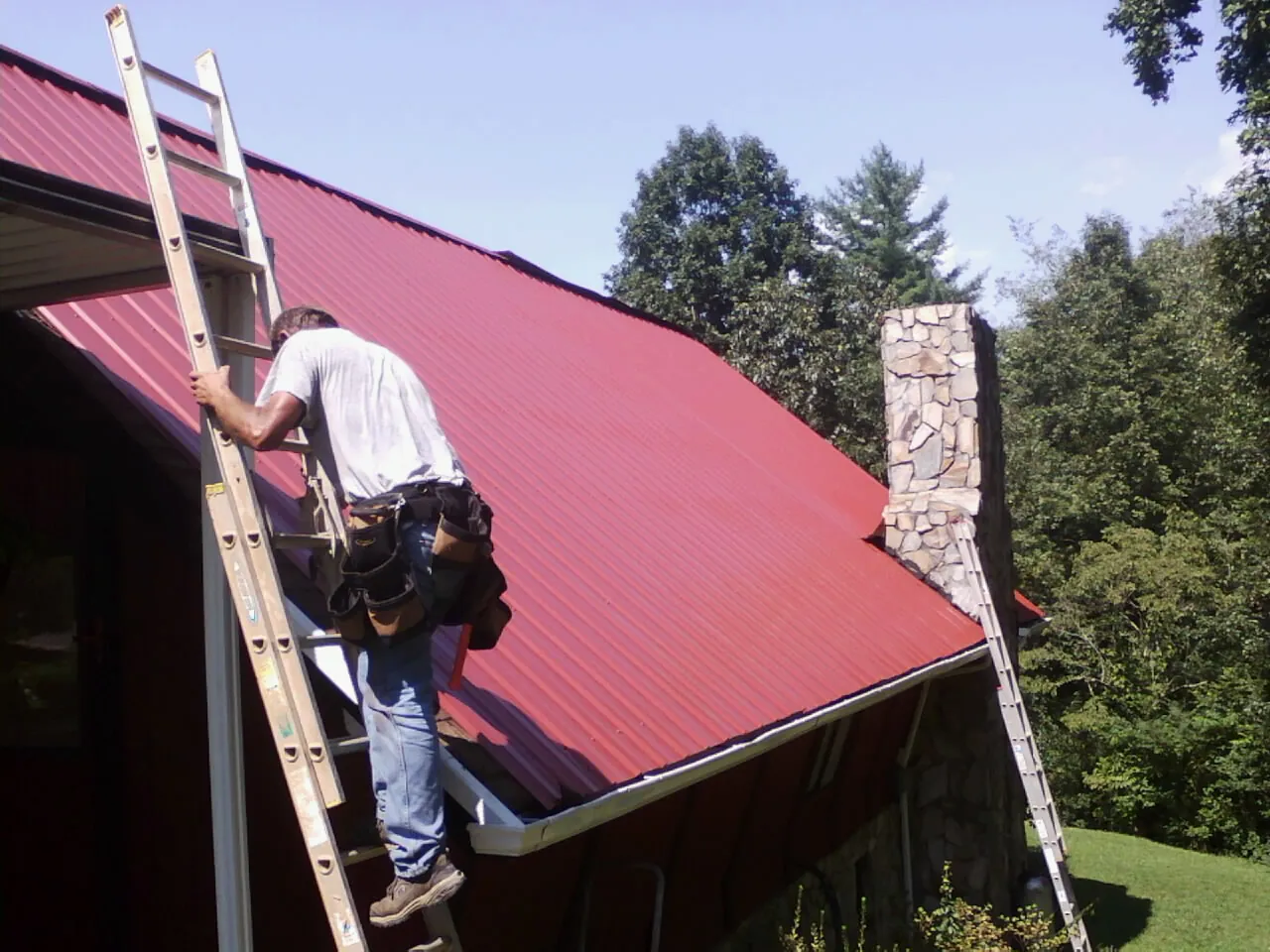 Roofer on a ladder beside a red metal roof and stone chimney — Soffit & Fascia Repair by Roofing Brothers in Conesus